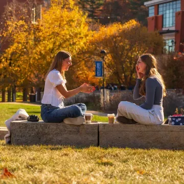 two students outside on the mall in the sunshine, fall, seated on bench